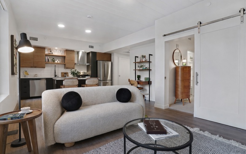 A living room with a couch, a center table, a side table, and views of a kitchen with a kitchen island countertop and barstool chairs, and stainless steel appliances at The Source on South Temple luxury apartments in Salt Lake City, UT.